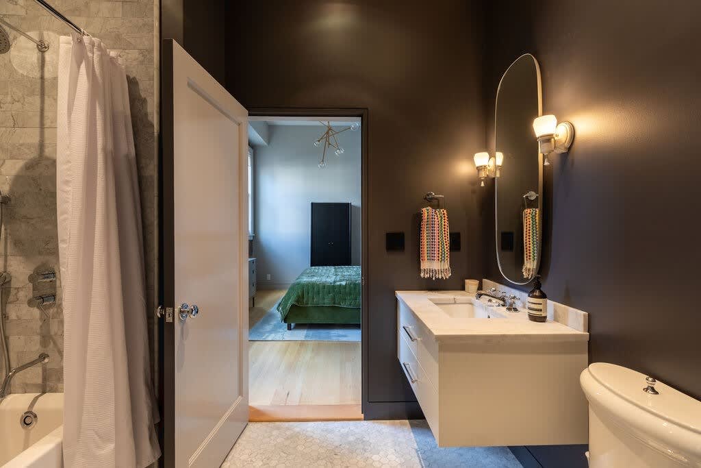 Compact bathroom with a white floating vanity and sink, an oval mirror with sconces, and a toilet. The floor is covered in small hexagonal tiles, and a shower/tub combo is visible behind an open door.