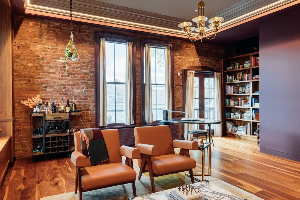 Cozy living room featuring exposed brick walls, two tan leather armchairs, a stylish bar cart, and a built-in bookshelf, all under a decorative ceiling with a chandelier.