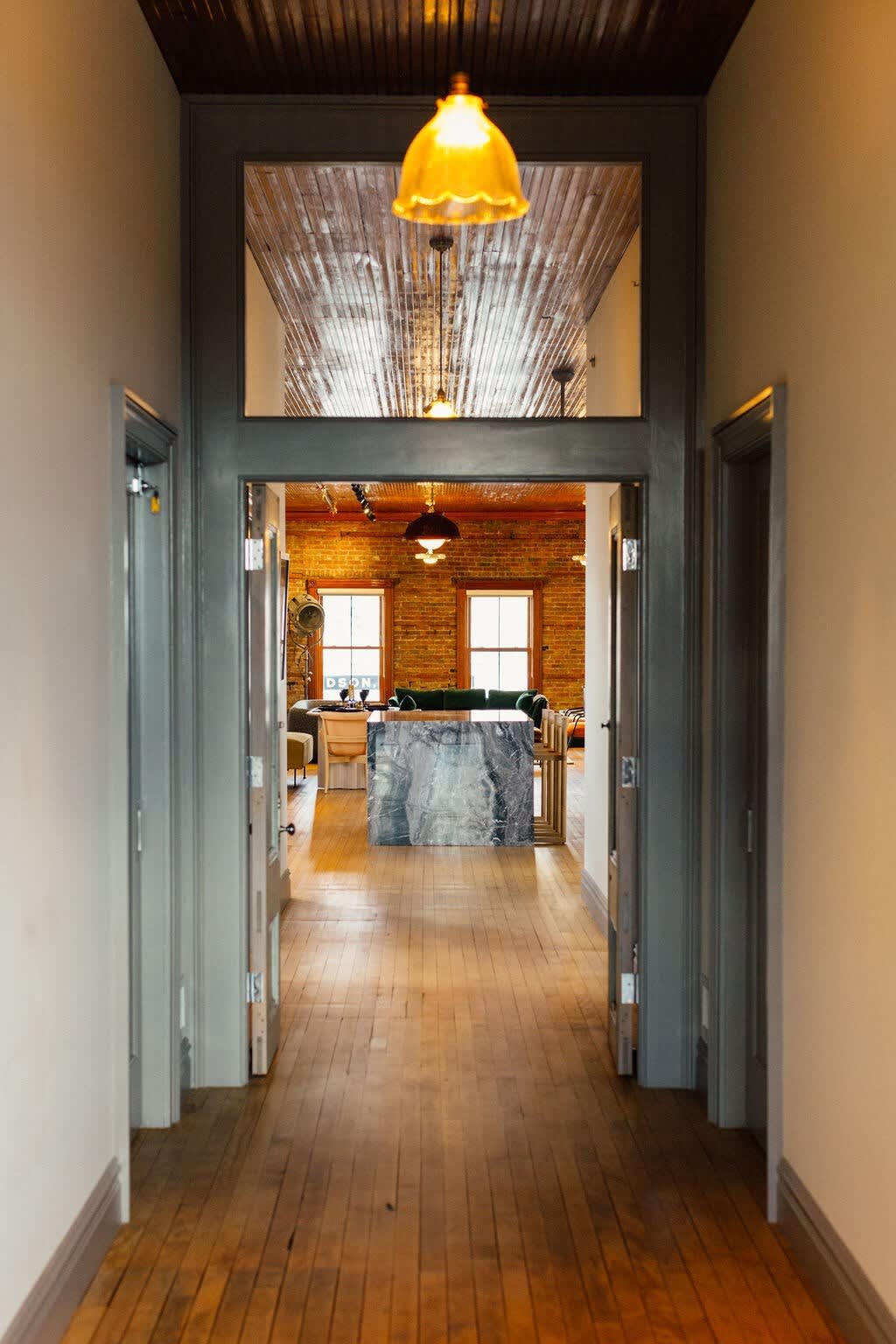Narrow hallway with light hardwood floors and gray trim, leading into a bright open living space with exposed brick walls and a warm wooden plank ceiling.