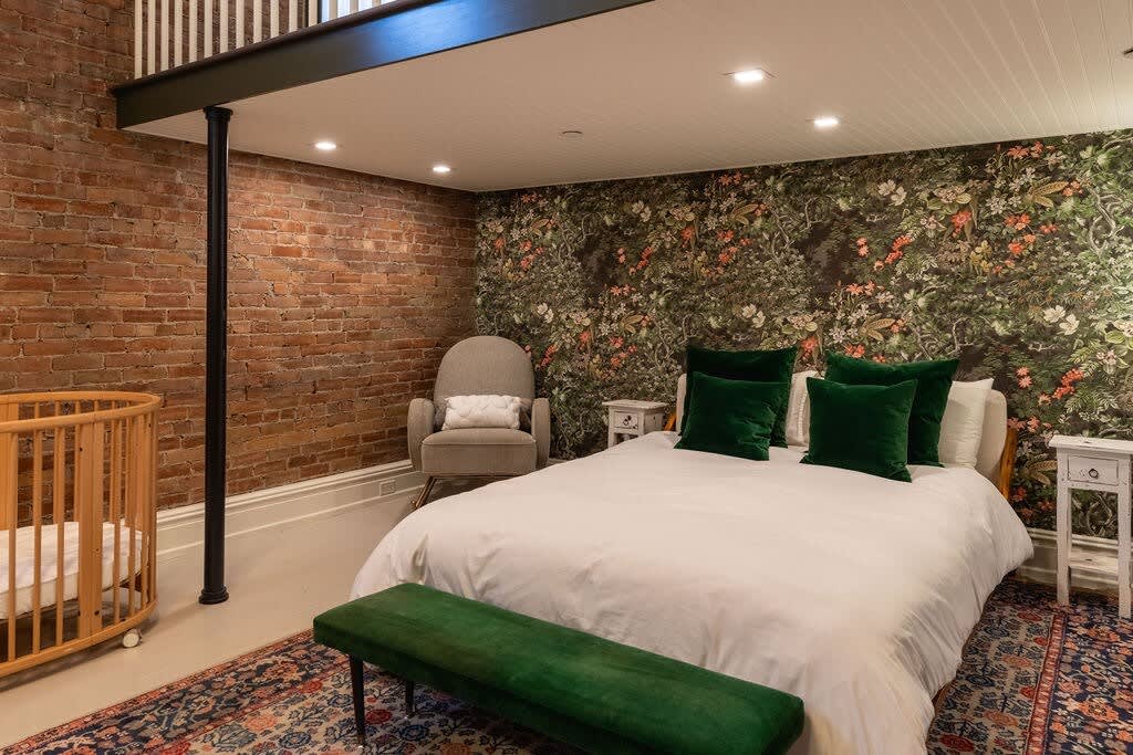 Cozy bedroom with a queen-sized bed featuring a white duvet and green velvet pillows, set against a dark floral wallpaper accent wall. A wooden crib and a gray rocking chair are visible, with exposed brick on the adjacent wall.