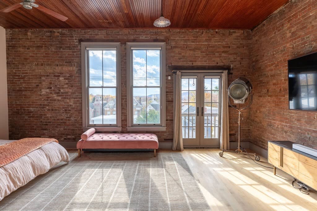 Wide view of a bright bedroom featuring an exposed brick wall, a pink chaise lounge, a large area rug, and French doors leading to an outdoor space, with a large TV on a wooden console.