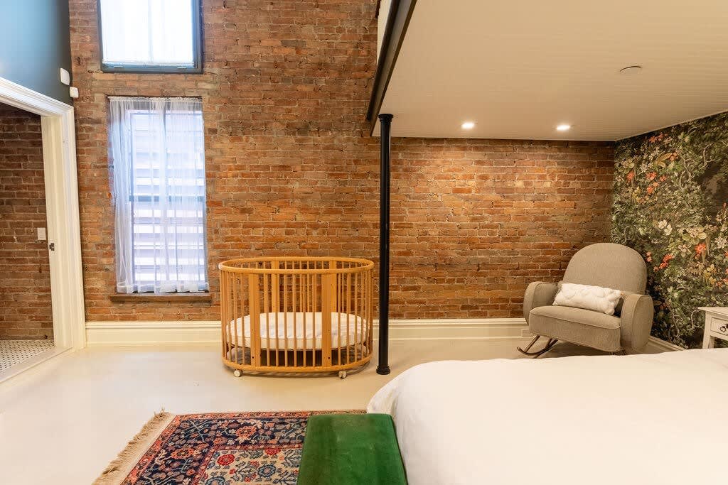 Bedroom with a wooden crib positioned against an exposed brick wall, next to a large window with sheer curtains. A gray rocking chair and part of a queen bed with a green velvet bench are also visible.