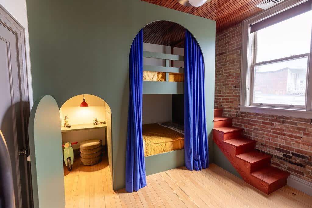 Playful children's bedroom featuring built-in green bunk beds with blue curtains and a red wooden staircase, alongside a whimsical arched nook with a desk and a small bike.