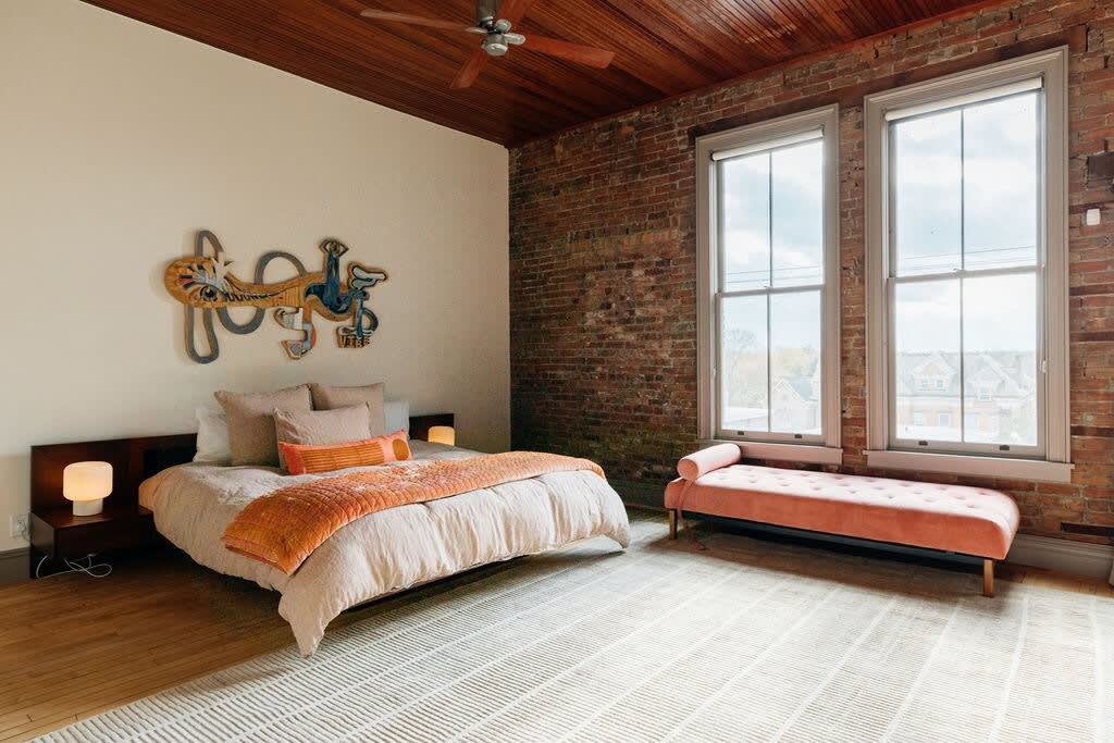 Spacious bedroom with a queen-sized bed adorned with orange and beige bedding, an exposed brick accent wall, and large windows, complemented by a rich wooden ceiling.