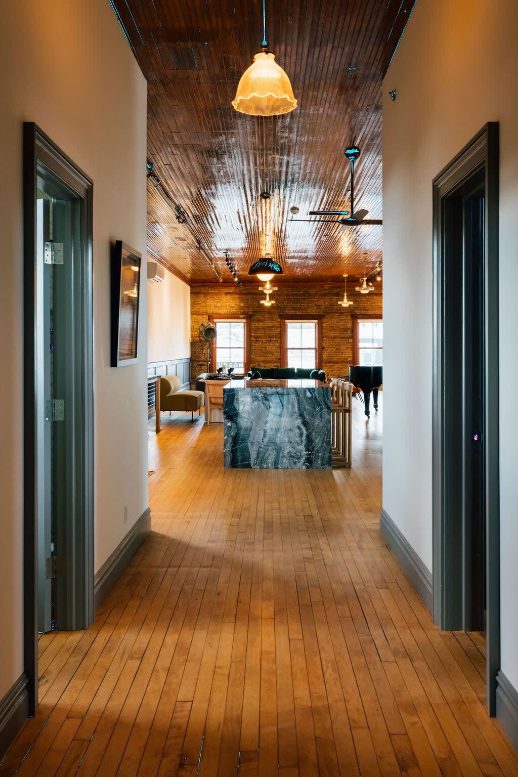 Long hallway with light hardwood floors and white walls, leading into an open-concept living area with exposed brick, a large marble island, and a rich wooden ceiling.