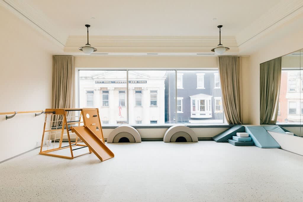 A spacious children's play area with a large window overlooking a city street, featuring a wooden climbing structure with a slide, soft play arches, and blue foam climbing blocks on a light carpeted floor.