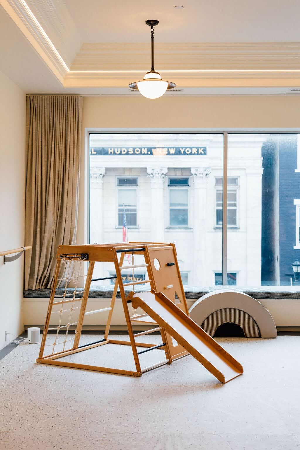 A bright children's play area with a large window offering city views, showcasing a wooden climbing structure with a slide, a ballet barre, and soft play arches on a light, textured floor.