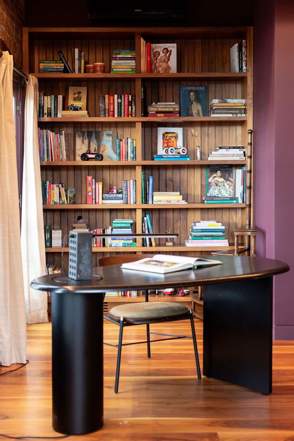 Dedicated office space with a dark oval desk and chair, positioned in front of a tall wooden bookshelf filled with books, set against a deep purple wall.