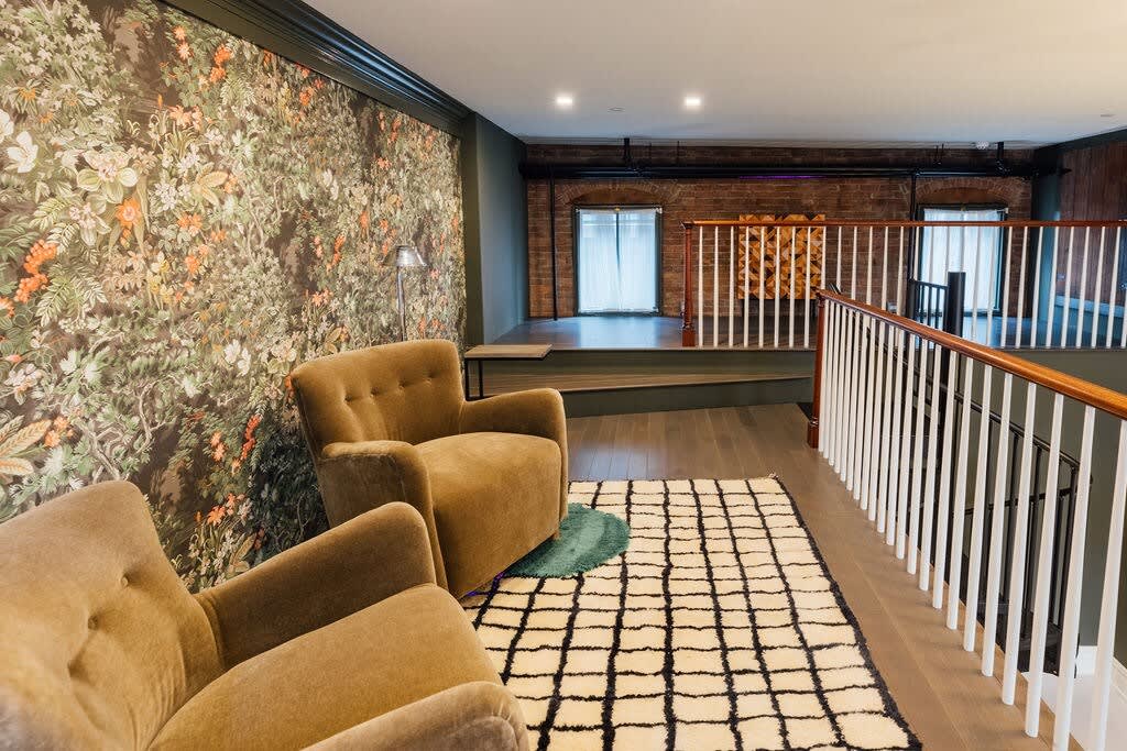 Inviting lounge area with two plush brown armchairs on a black and white checkered rug, set against a dark floral wallpaper accent wall. A white railing separates the space from a lower level, with exposed brick and windows in the background.