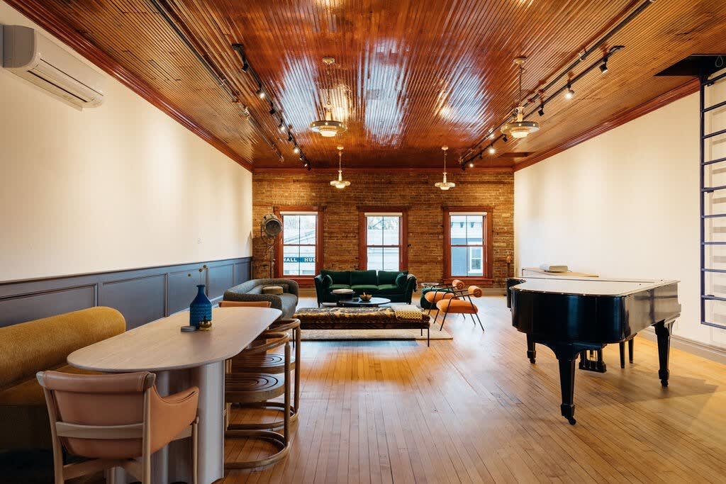 Expansive open-plan living area with a grand piano, a green velvet sofa, and a dining area with a long wooden table, all under a glossy wooden ceiling with industrial lighting.