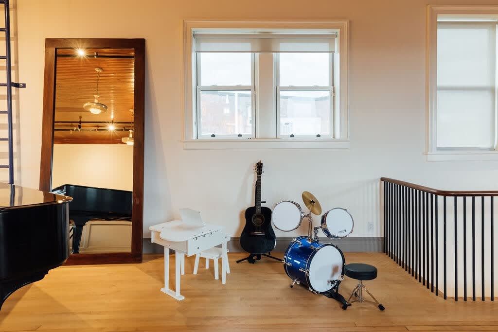 Bright open space featuring a grand piano, a guitar, and a blue drum set, with large windows and a full-length mirror reflecting the wooden ceiling.