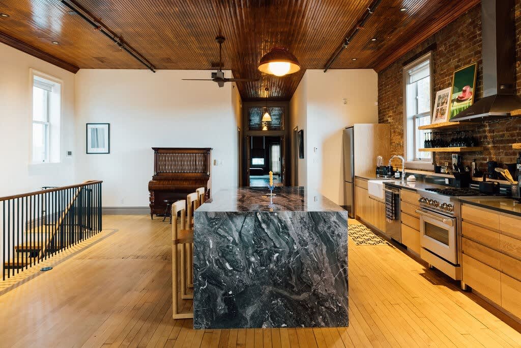 Spacious kitchen and dining area featuring a large black marble island, light wood cabinetry, exposed brick walls, and a warm wooden plank ceiling, with a view of a grand piano.
