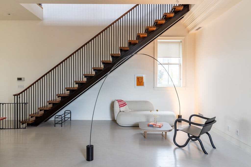 Modern living area with a striking open-tread staircase, a minimalist white sofa, a unique black armchair, and an arched floor lamp, all against a white wall.