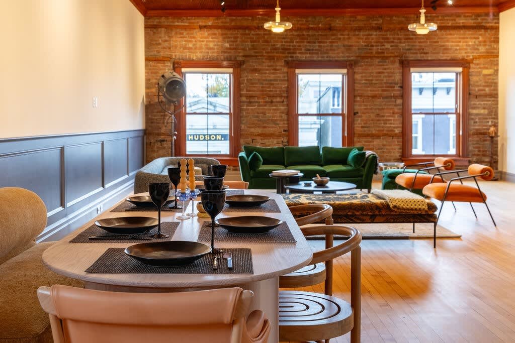 Open-concept dining area with a light wood table set for dinner, leading into a vibrant living space with exposed brick walls, a green sofa, and orange armchairs.