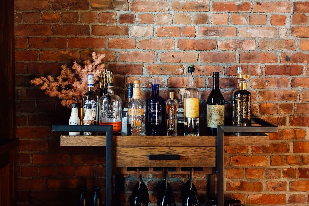 Close-up of a rustic wooden bar cart stocked with various liquor bottles and wine glasses, set against an exposed brick wall.