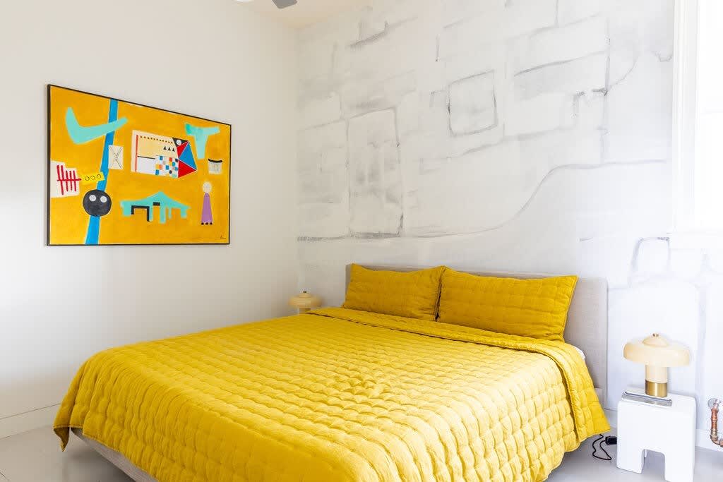 A cozy bedroom with a yellow quilted bed, an abstract painting on the wall, and a textured white accent wall, featuring a modern bedside lamp and a light-colored headboard.