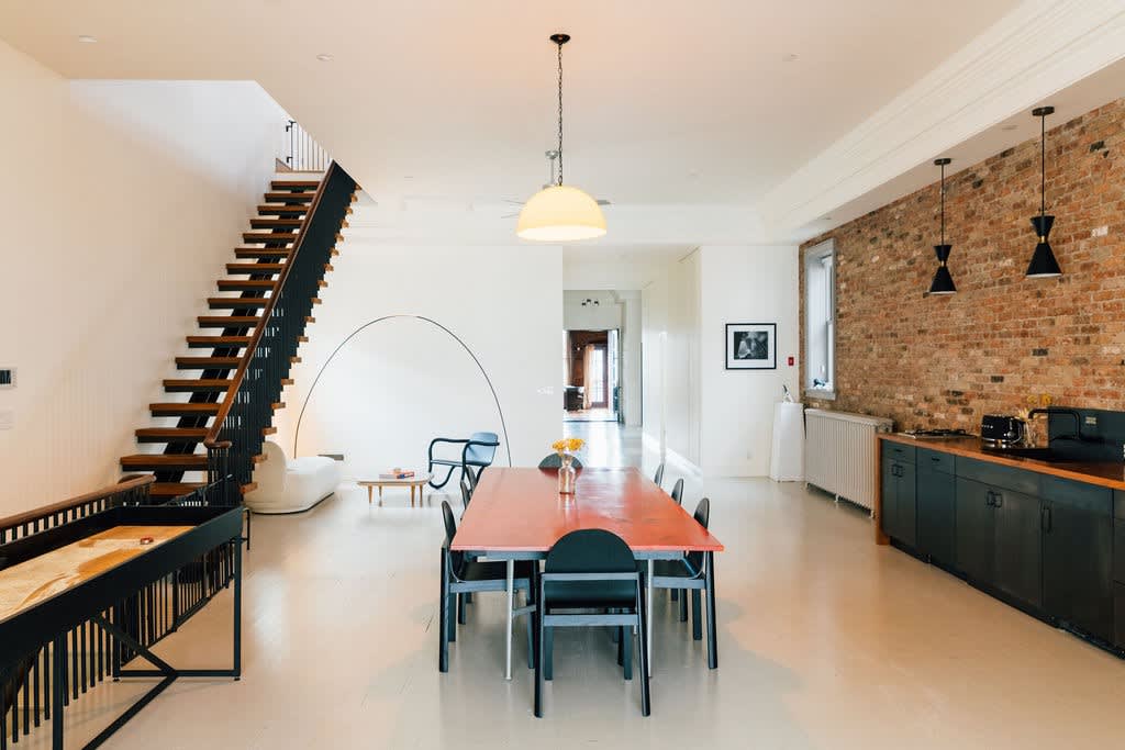An expansive open-concept living space with a long orange dining table, a shuffleboard table, a modern staircase, and exposed brick walls, illuminated by a large pendant light and recessed lighting.
