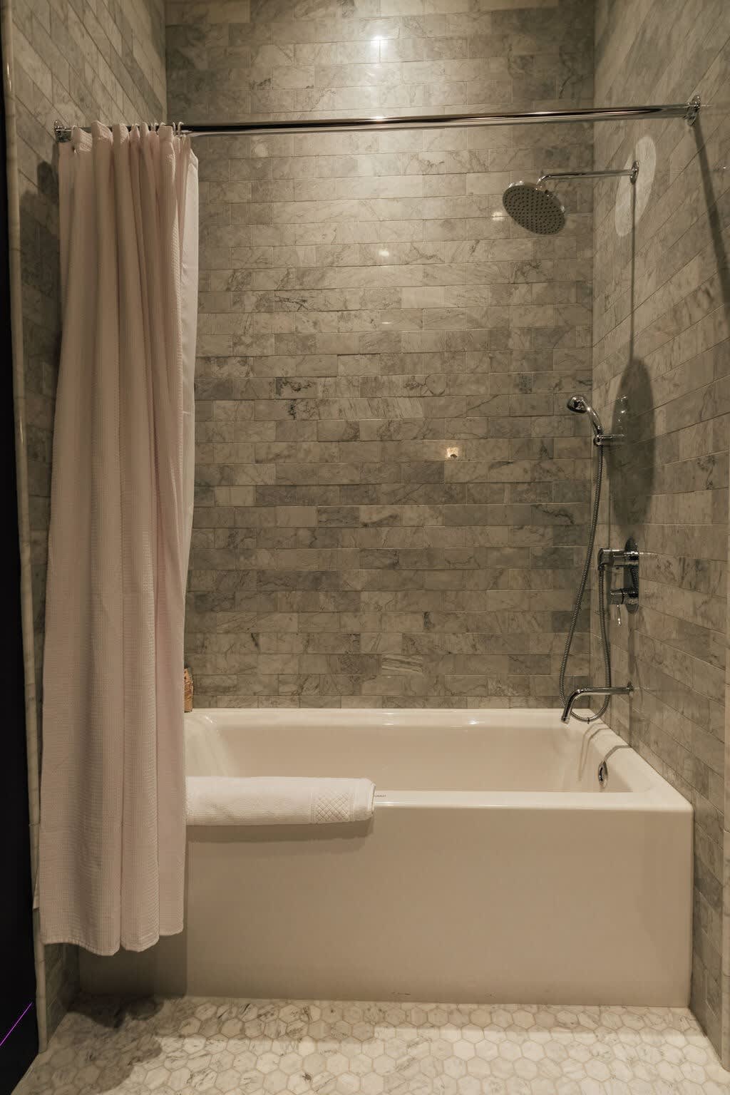Bathroom featuring a white bathtub with a shower curtain, surrounded by light gray rectangular subway tiles on the walls. The floor has small hexagonal tiles, and a shower head and faucet are visible.