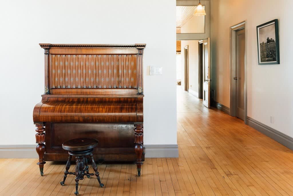 Bright hallway with polished light wood floors and white walls, featuring an antique wooden upright piano with a matching stool. A doorway leads to another room, and a framed picture hangs on the wall.