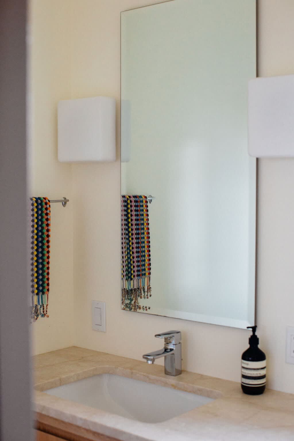 A close-up of a modern bathroom vanity with a light stone countertop, an integrated sink, a sleek faucet, a large rectangular mirror, and wall-mounted sconces, with colorful towels hanging nearby.