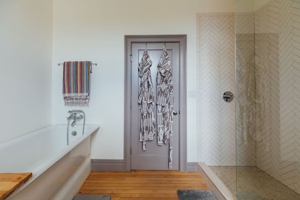 Elegant bathroom featuring a freestanding bathtub, a glass-enclosed walk-in shower with herringbone tile, and light hardwood floors, with patterned robes hanging on the door.