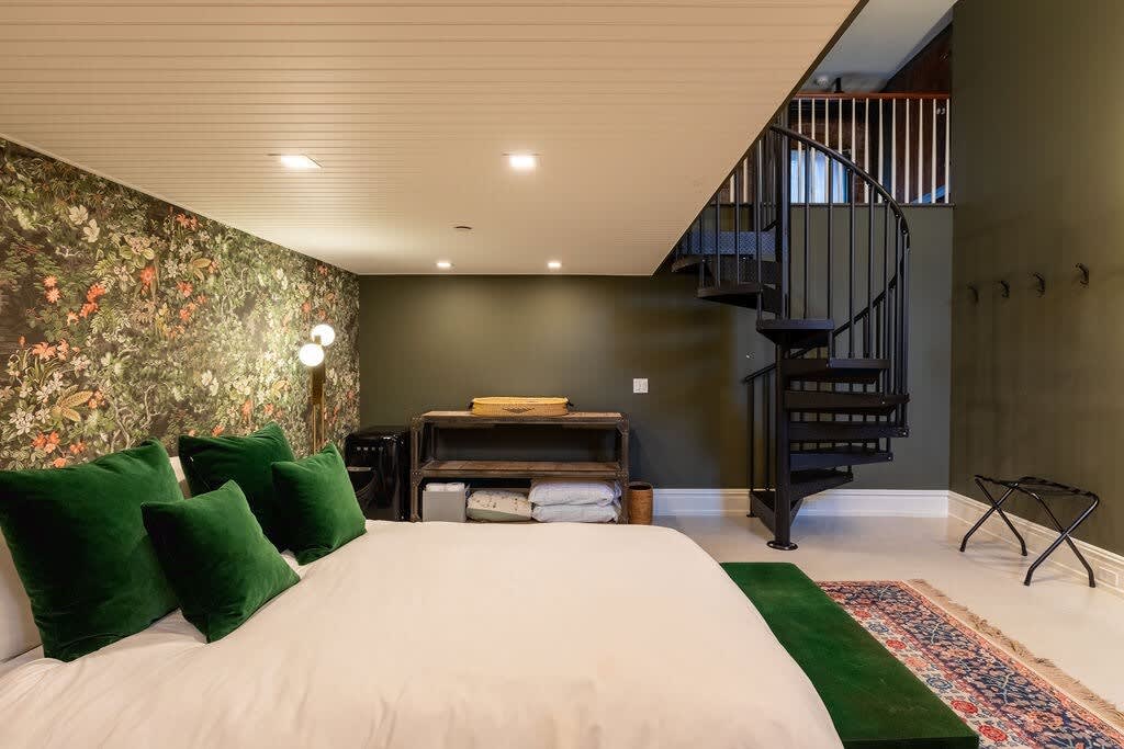 Bedroom featuring a queen bed with green velvet pillows, a dark floral accent wall, and a black spiral staircase leading to an upper level. A wooden shelving unit and a luggage rack are also visible.