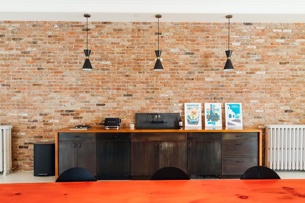 A dining area featuring a long orange table with black chairs, set against an exposed brick wall with a dark wood counter below, adorned with three modern pendant lights and various kitchen appliances.