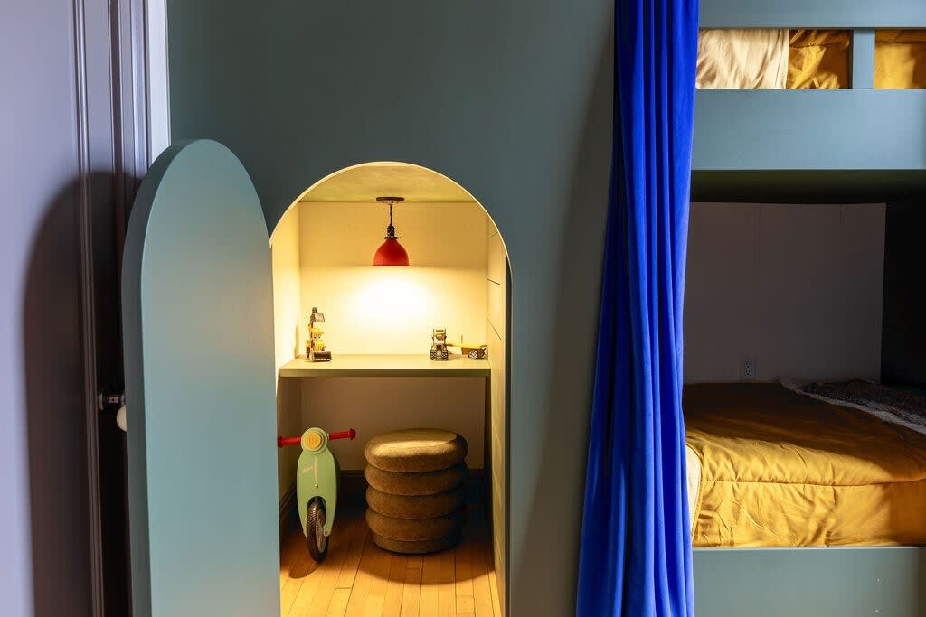 Close-up of a charming arched play nook in a children's bedroom, featuring a small desk with toys, a red pendant light, and a miniature bike, all within a sage green built-in structure.