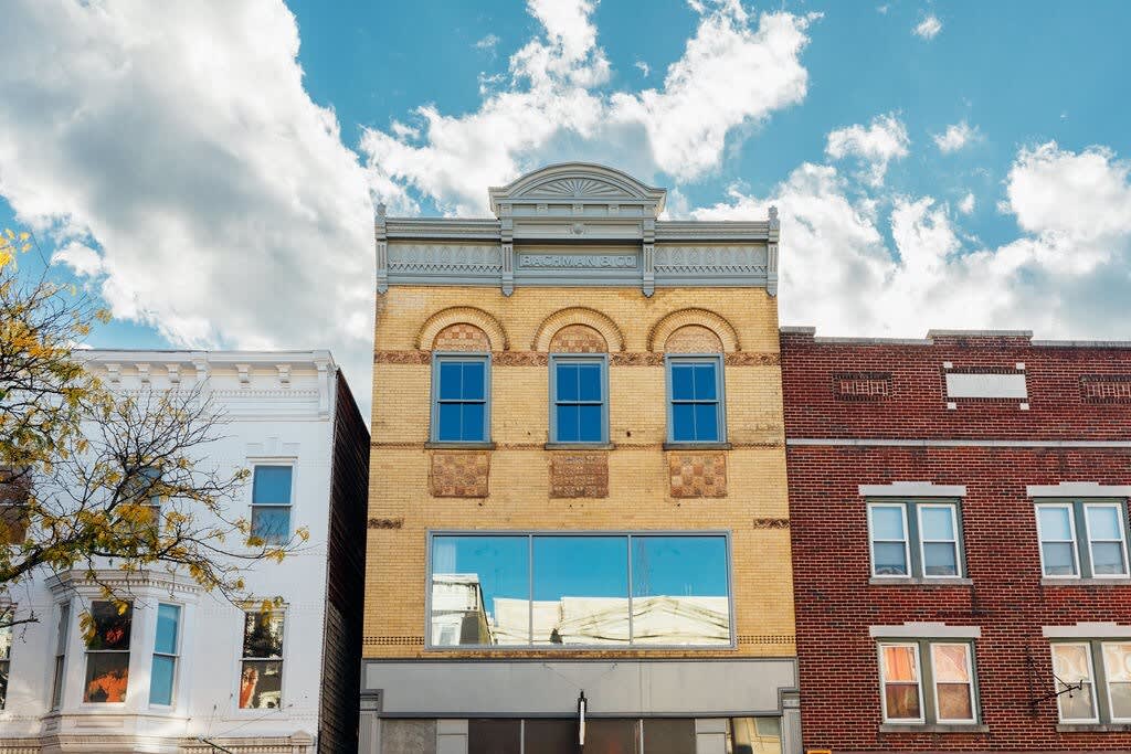 A vibrant street view showcasing three distinct buildings under a bright blue sky with fluffy white clouds. The central building features a yellow brick facade with arched windows and a decorative gray cornice, flanked by a white building on the left and a red brick building on the right.