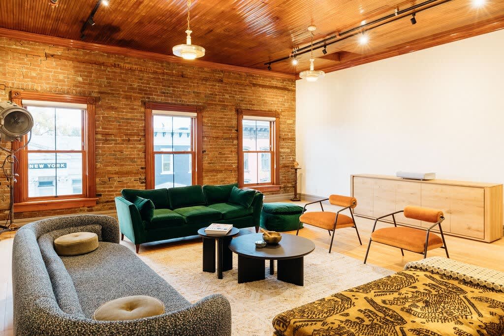 Expansive living area with exposed brick walls, a rich wooden ceiling, a green velvet sofa, a grey textured sofa, and two orange armchairs arranged around dark coffee tables on a light rug.