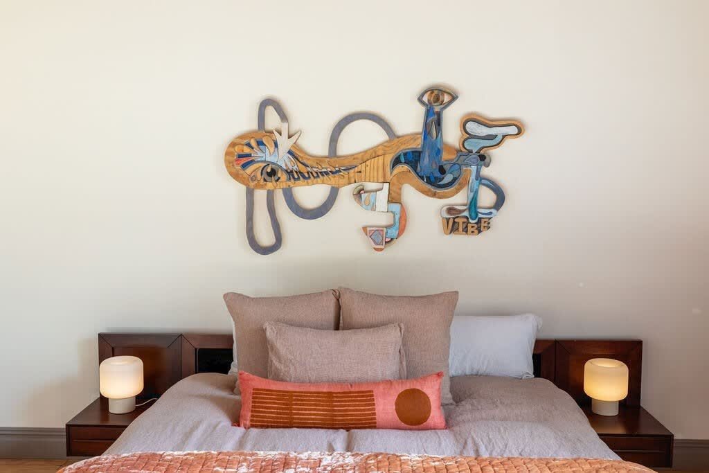 Close-up of a queen-sized bed with light pink and orange bedding, flanked by modern bedside tables with glowing lamps, and a unique abstract art piece mounted above the headboard.