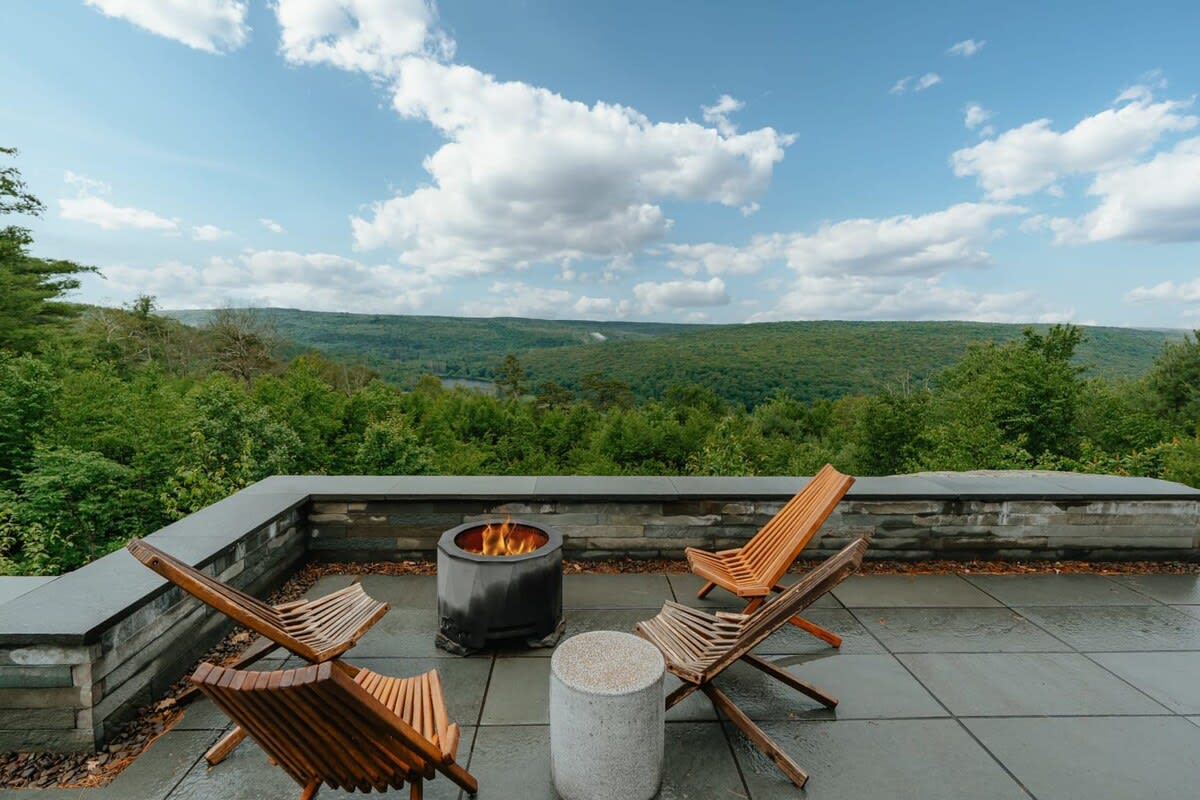 Outdoor patio with a stone-clad wall and large square pavers, featuring a circular fire pit surrounded by four slatted wooden Adirondack-style chairs, offering expansive views of a green, rolling forested valley under a cloudy sky.