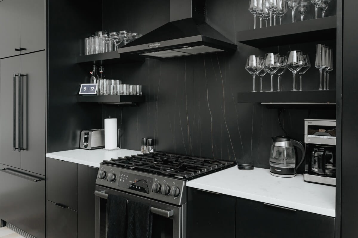 Detailed view of the modern kitchen's cooking area, featuring a gas range, white countertops, and black open shelves filled with glassware. A toaster and coffee maker are also visible, highlighting functional amenities.