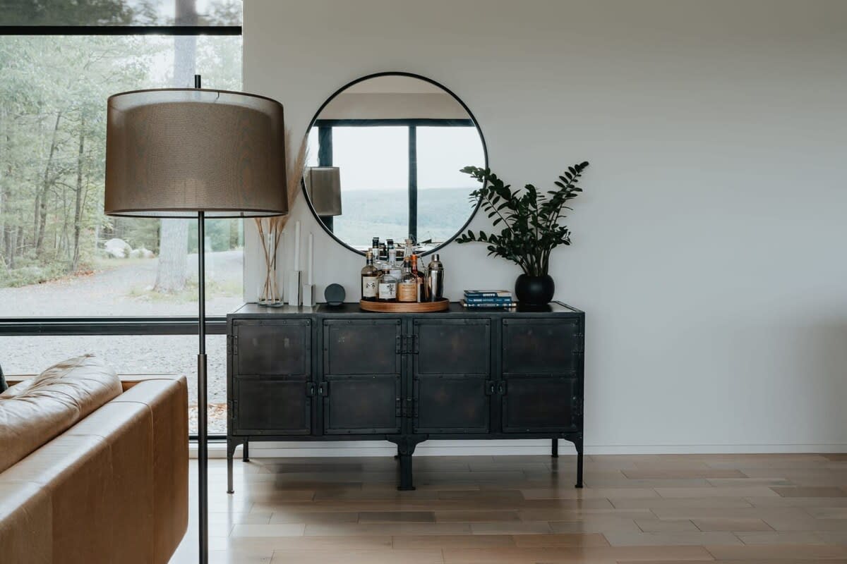 A modern industrial-style black metal cabinet with a bar setup on top, featuring various liquor bottles and bar tools, stands against a white wall with a large round mirror reflecting the outdoors. A tall floor lamp and a brown leather sofa are partially visible on the left.