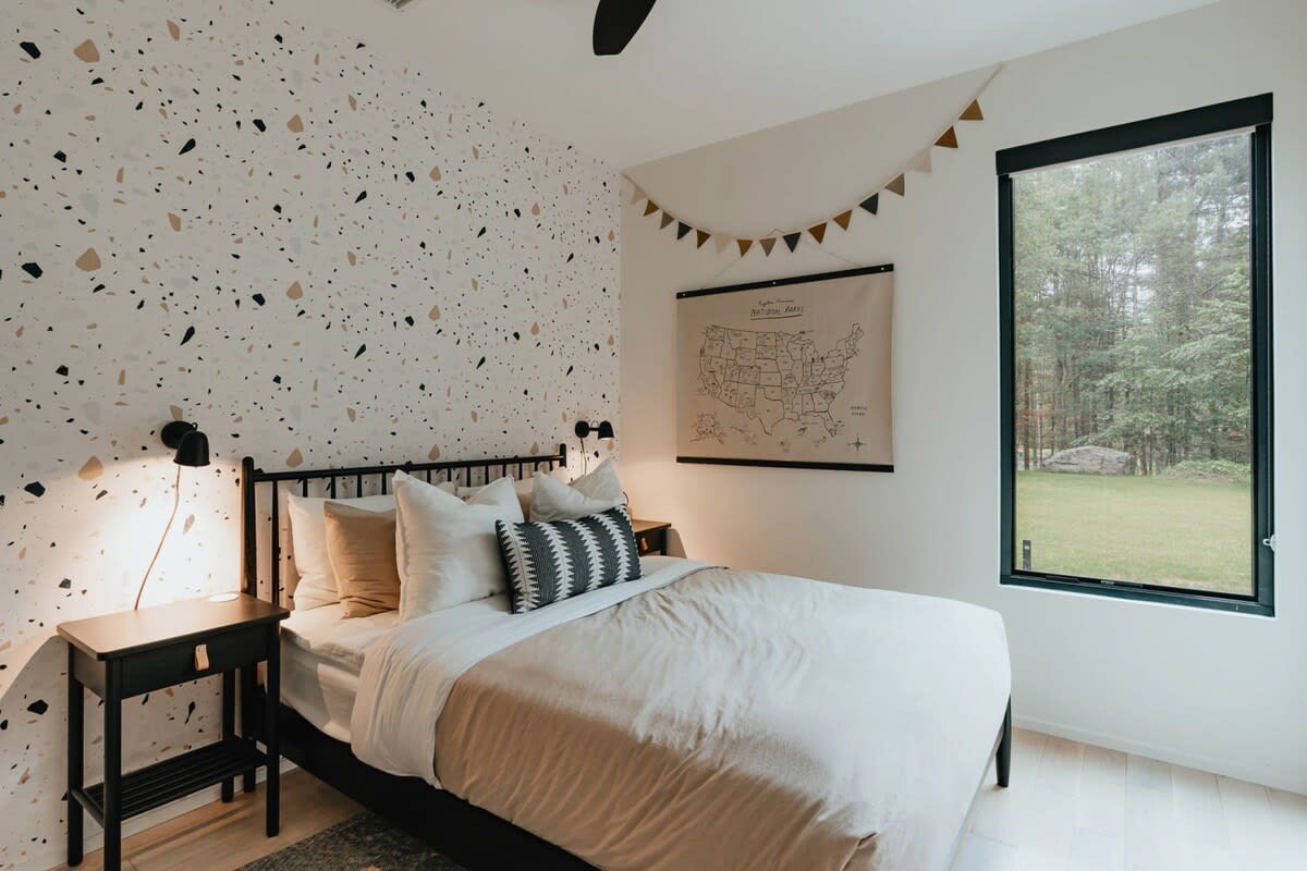 A cozy bedroom features a black metal bed frame with light bedding and a patterned throw pillow, set against a white wall with a playful terrazzo-patterned accent wall. A black nightstand with a lamp and a large window overlooking a wooded area are visible.