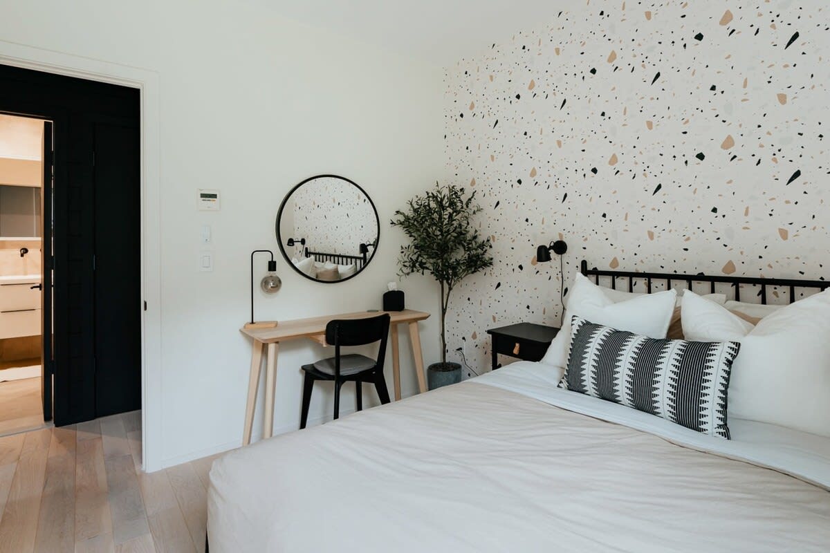 This bedroom features a light wood desk with a black chair and a round mirror, positioned against a white wall adjacent to a terrazzo-patterned accent wall. A potted plant adds greenery, and a black door leads to an en-suite bathroom.