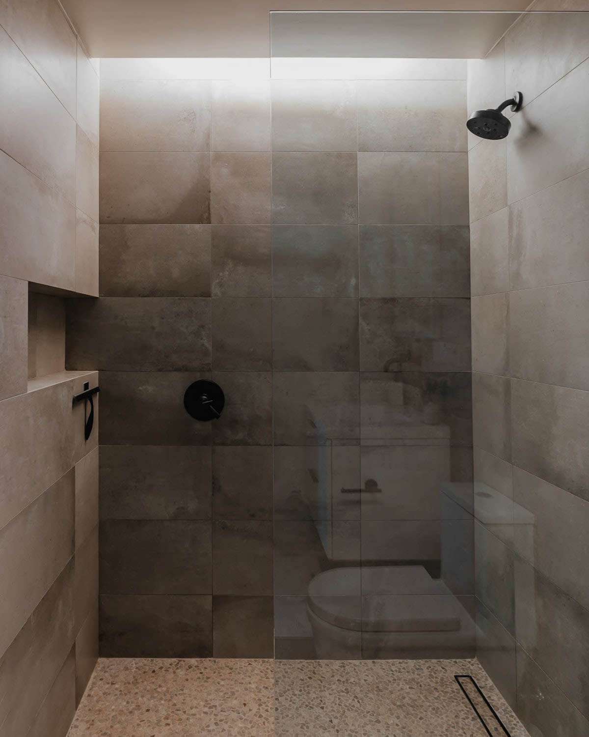 A close-up view inside a modern walk-in shower with light grey tiled walls and a speckled terrazzo floor. It features a black shower head, a built-in niche, and a glass partition, with the toilet visible through the glass.