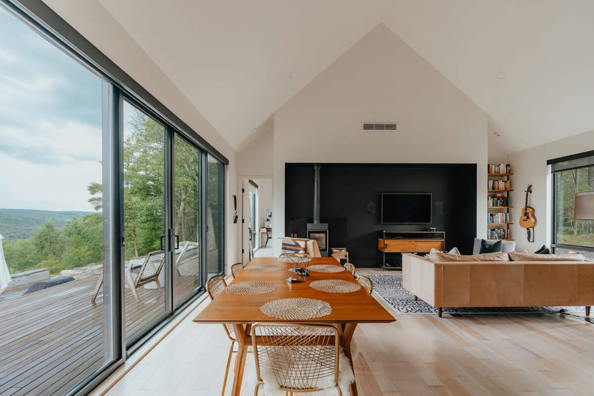 Expansive open-concept living and dining area with a vaulted ceiling, featuring a wooden dining table, a cozy living space with a modern fireplace, and floor-to-ceiling sliding glass doors leading to an outdoor deck with forest views.