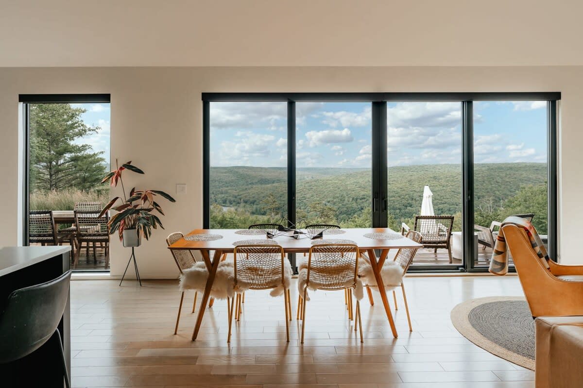Bright dining area with a long wooden table and six woven chairs, set against a backdrop of floor-to-ceiling sliding glass doors that offer panoramic views of a lush green forest and distant hills.