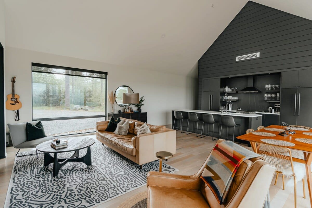 Open-concept living space with a vaulted ceiling, combining a living room, dining area, and a modern black kitchen. Large windows offer expansive views of the surrounding nature.