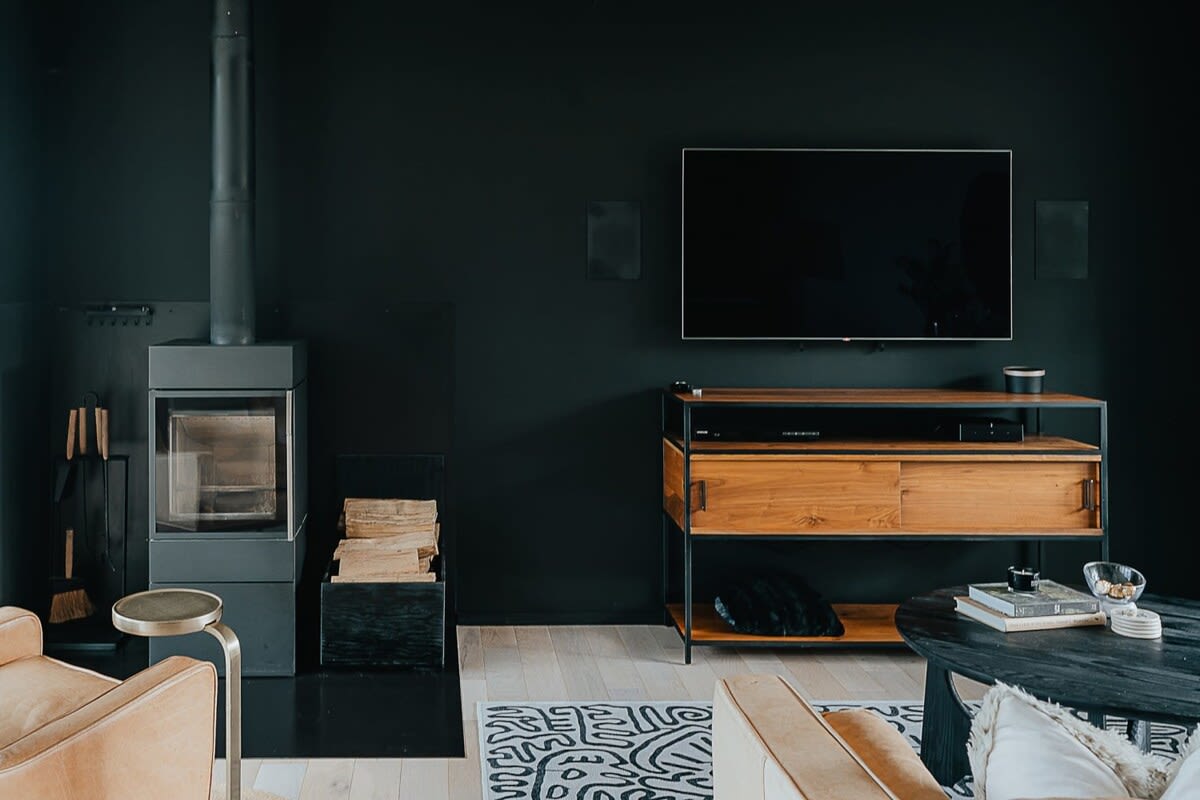 Close-up of the living room's black accent wall featuring a modern wood-burning stove, a wall-mounted TV, and a wooden media console. Stacked firewood adds a rustic touch to the contemporary design.
