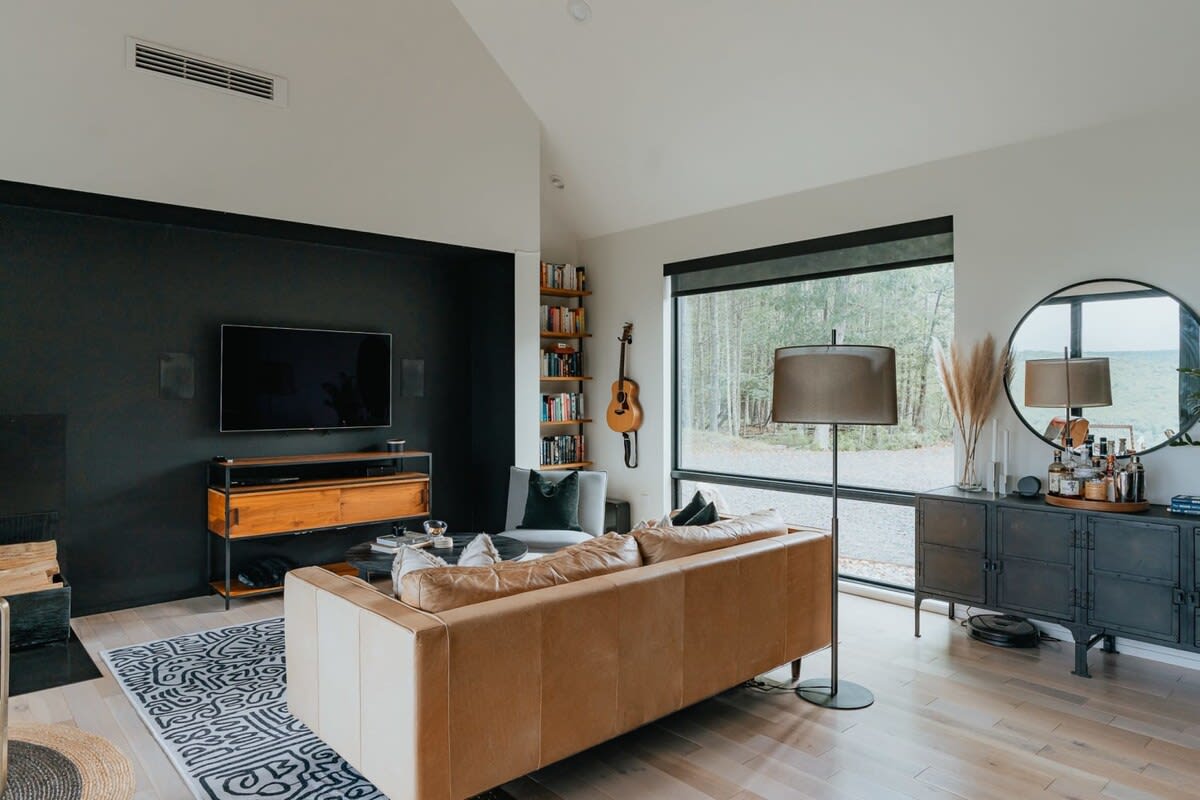 Spacious living room with a vaulted ceiling, light hardwood floors, and a large window overlooking the forest. A tan leather sofa, a black accent wall with a TV, and a bookshelf are prominent.
