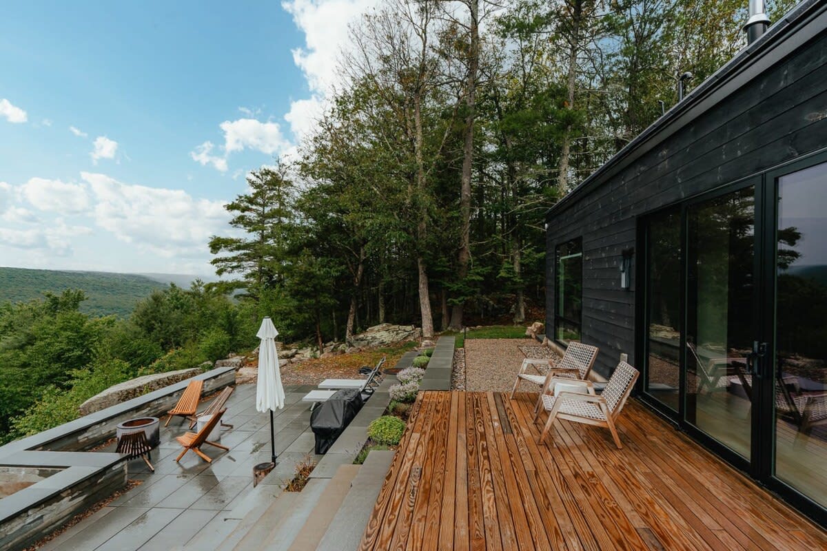 Wooden deck leading to a stone patio with outdoor seating and an umbrella, offering expansive views of a lush green valley and distant mountains under a blue sky.