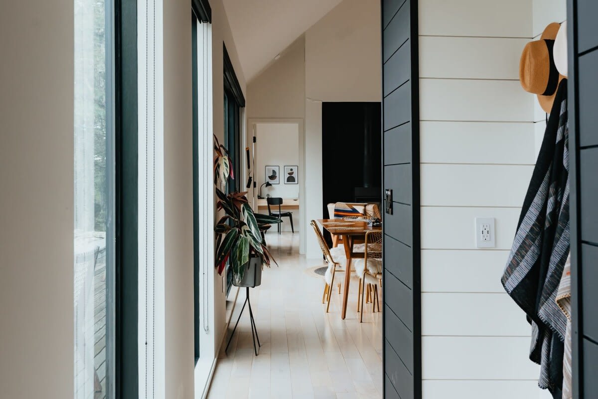 View from the entryway into an open-concept living space with light hardwood floors and large windows. A dining area and a plant on a stand are visible in the distance, leading to other rooms.