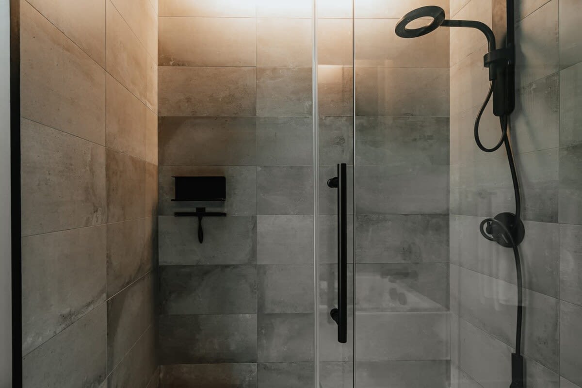 Walk-in shower with large grey and beige tiled walls, a glass door with a black handle, and a black rain shower head and handheld sprayer.