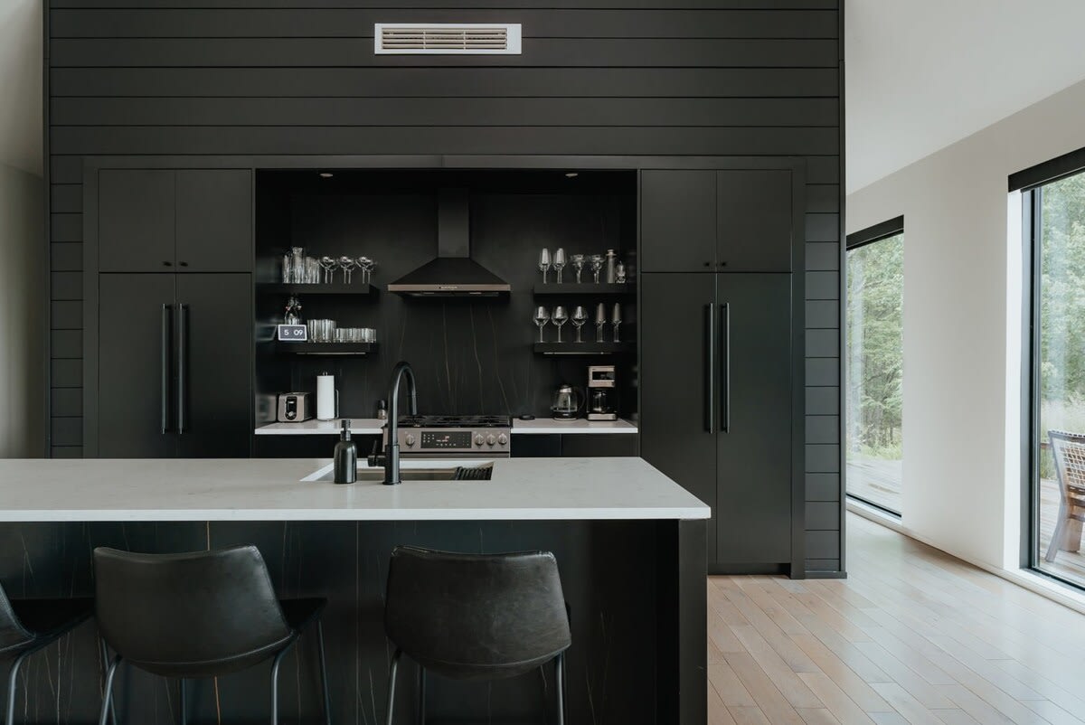 Sleek, modern kitchen with dark cabinetry, a large island with seating, and integrated appliances. Open shelving displays glassware, and a black range hood is centrally placed above the stove area.