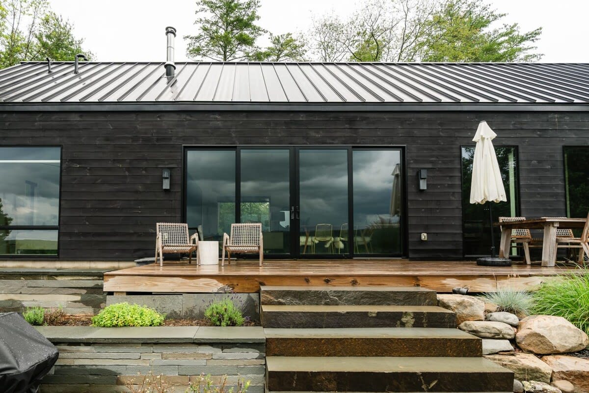 Rear exterior of the black modern house with large glass sliding doors opening onto a wooden deck, furnished with outdoor chairs and an umbrella, overlooking a landscaped area.