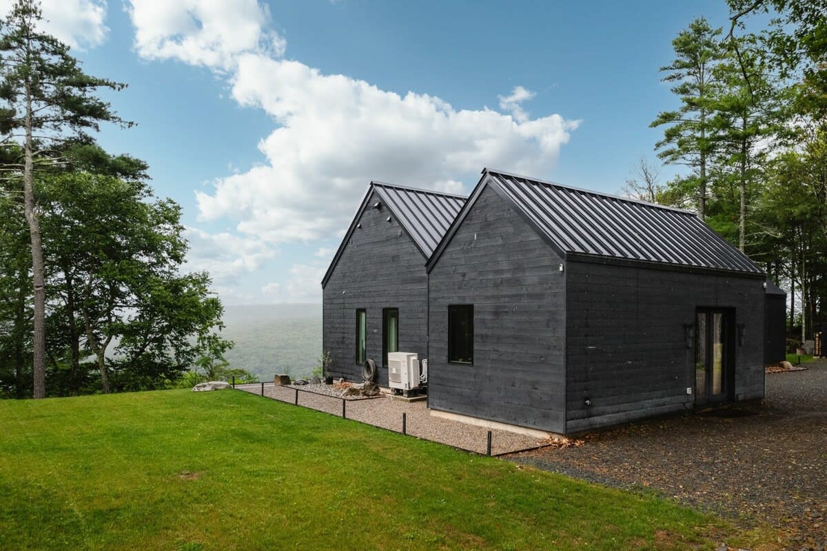 Side view of a modern black house with two gabled sections and dark wood siding, set on a grassy hill with a gravel path, surrounded by tall trees and offering distant scenic views.