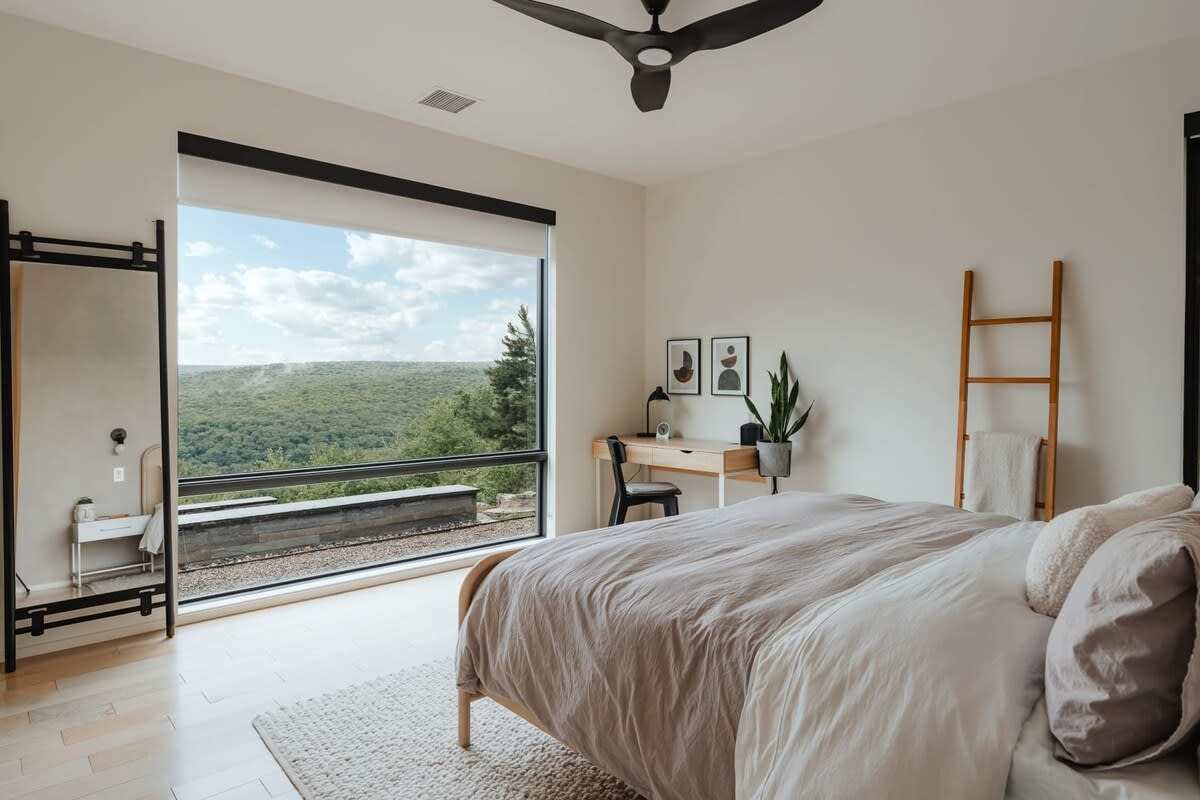 Modern bedroom with light walls, a large window offering panoramic forest views, and a light-colored bed with a ceiling fan. A desk and a decorative ladder are also visible.