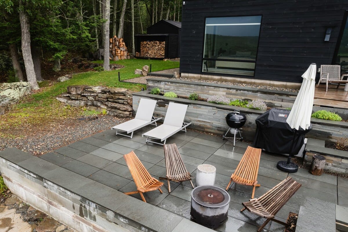 Multi-level outdoor patio featuring grey stone pavers, wooden lounge chairs, a barbecue grill, and a fire pit, surrounded by low landscaping and mature trees.